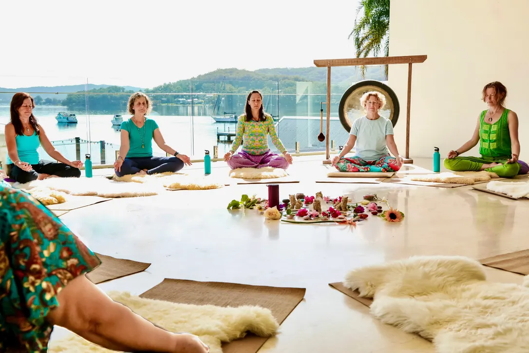 Women sit cross-legged on sheepskins in a circle around a flower mandala, a gong and the bay beyond the studio windows